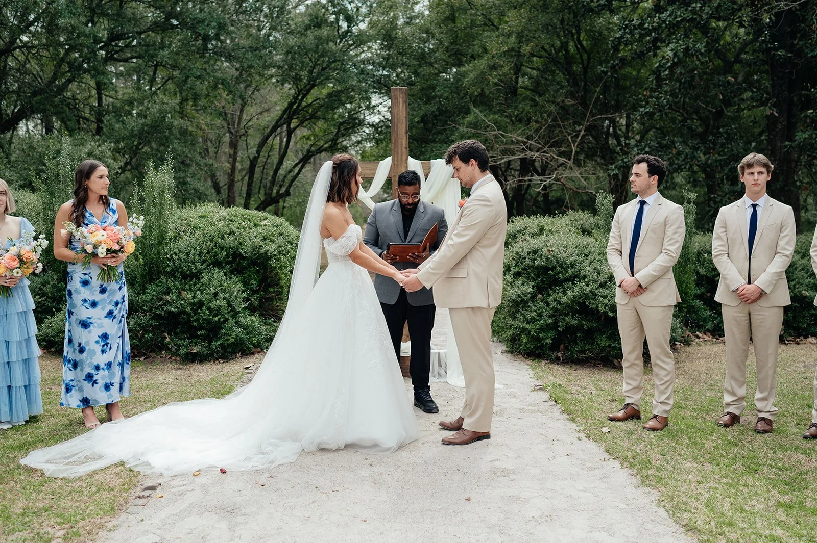 Bride and groom exchange vows in a garden ceremony surrounded by greenery and pastel florals, with bridesmaids and groomsmen standing nearby — coordinated by Charlotte Wedding Coordinator.