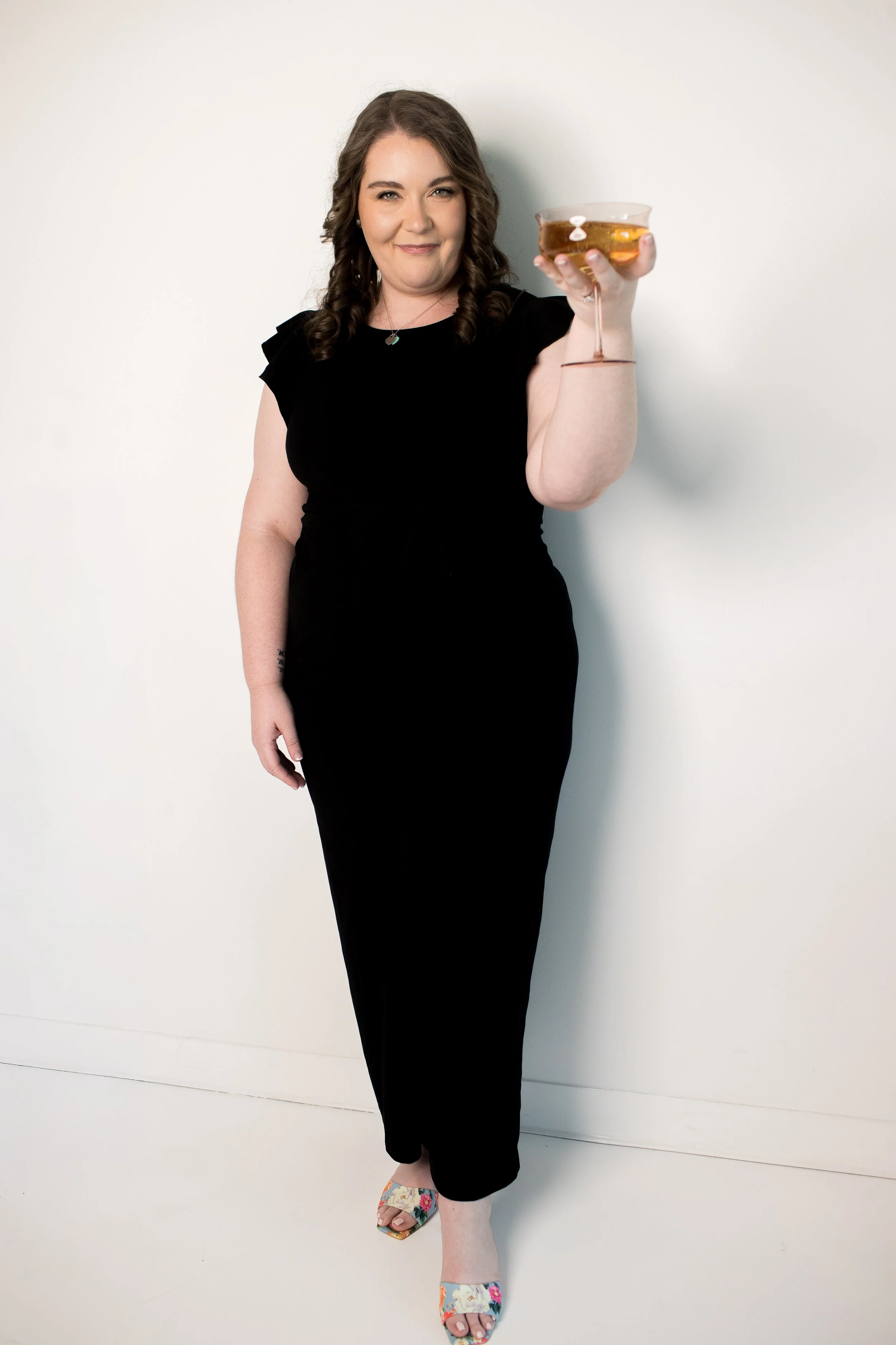 A Charlotte wedding planner holding a cocktail glass with a drink, standing against a plain white wall.