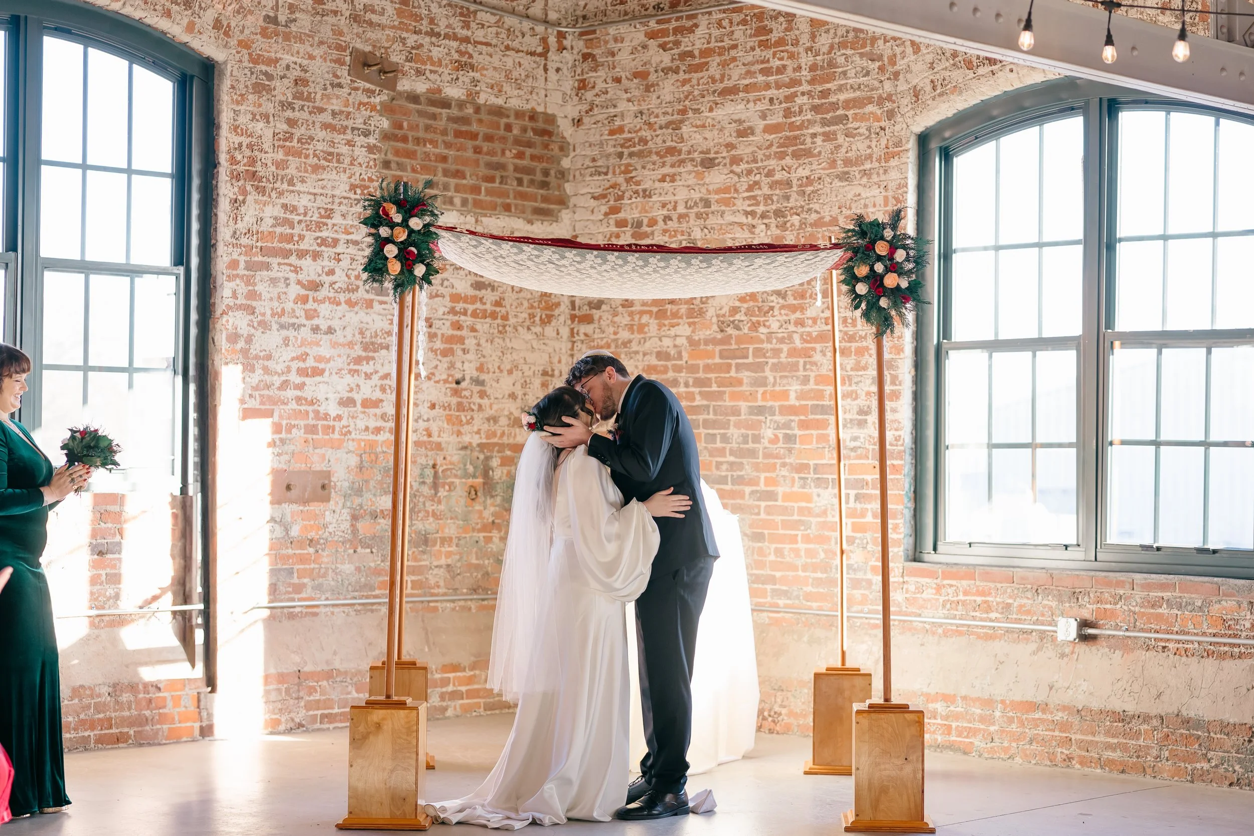 Bride and groom share a kiss beneath a floral canopy in a modern brick venue filled with natural light and romantic details — coordinated by Charlotte Wedding Coordinator.
