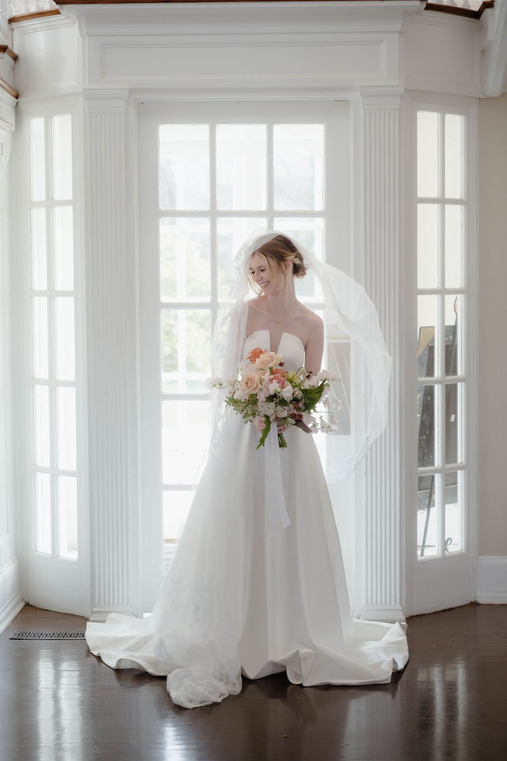 A bride in a strapless white wedding gown holding a bouquet of pink, peach, and white flowers stands in front of a large window with white frames, smiling.