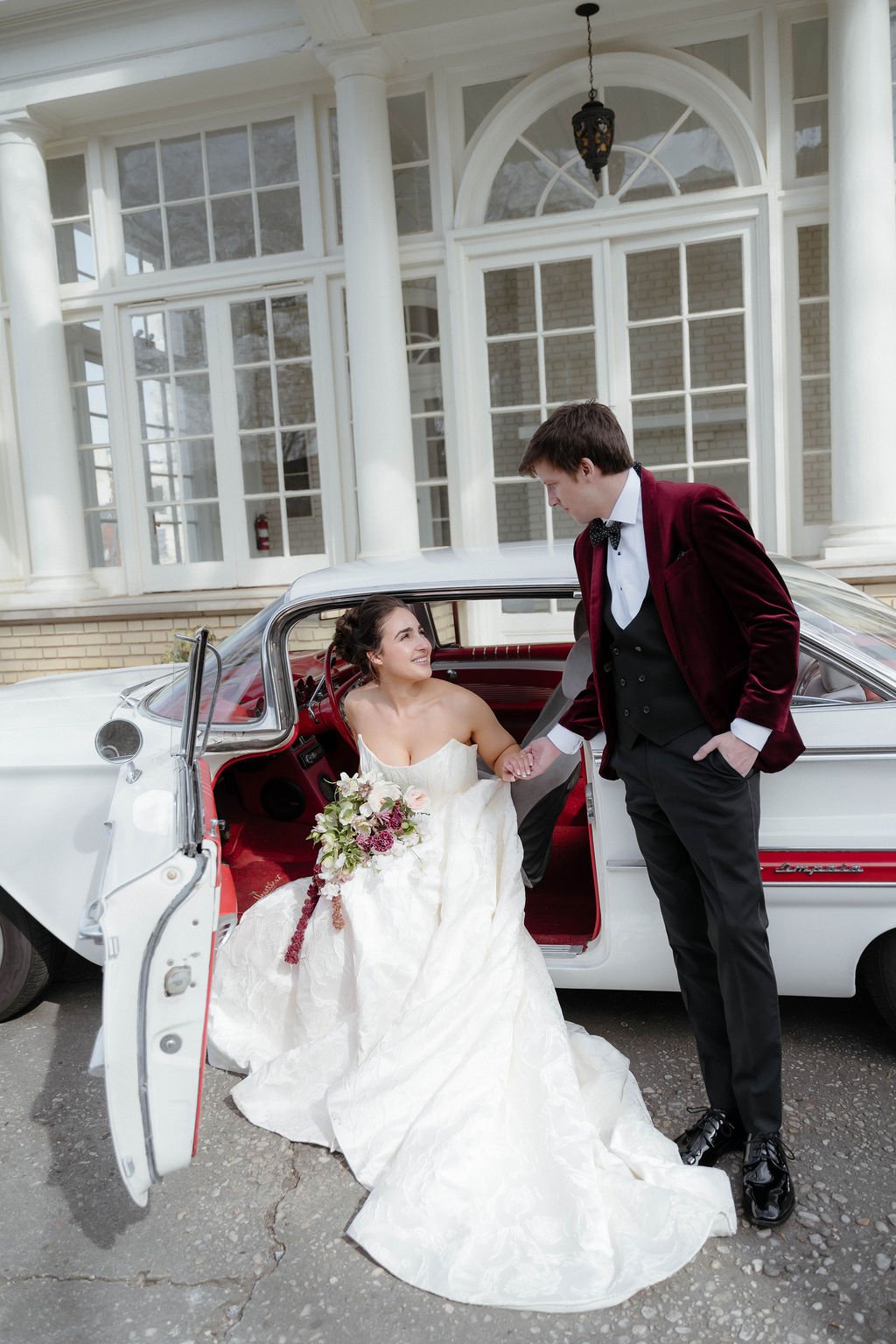 A bride in a white wedding gown holding a bouquet of flowers sitting inside a vintage white car, holding hands with a groom in a black tuxedo and burgundy velvet blazer outside the car, in front of a large building with white columns and windows.