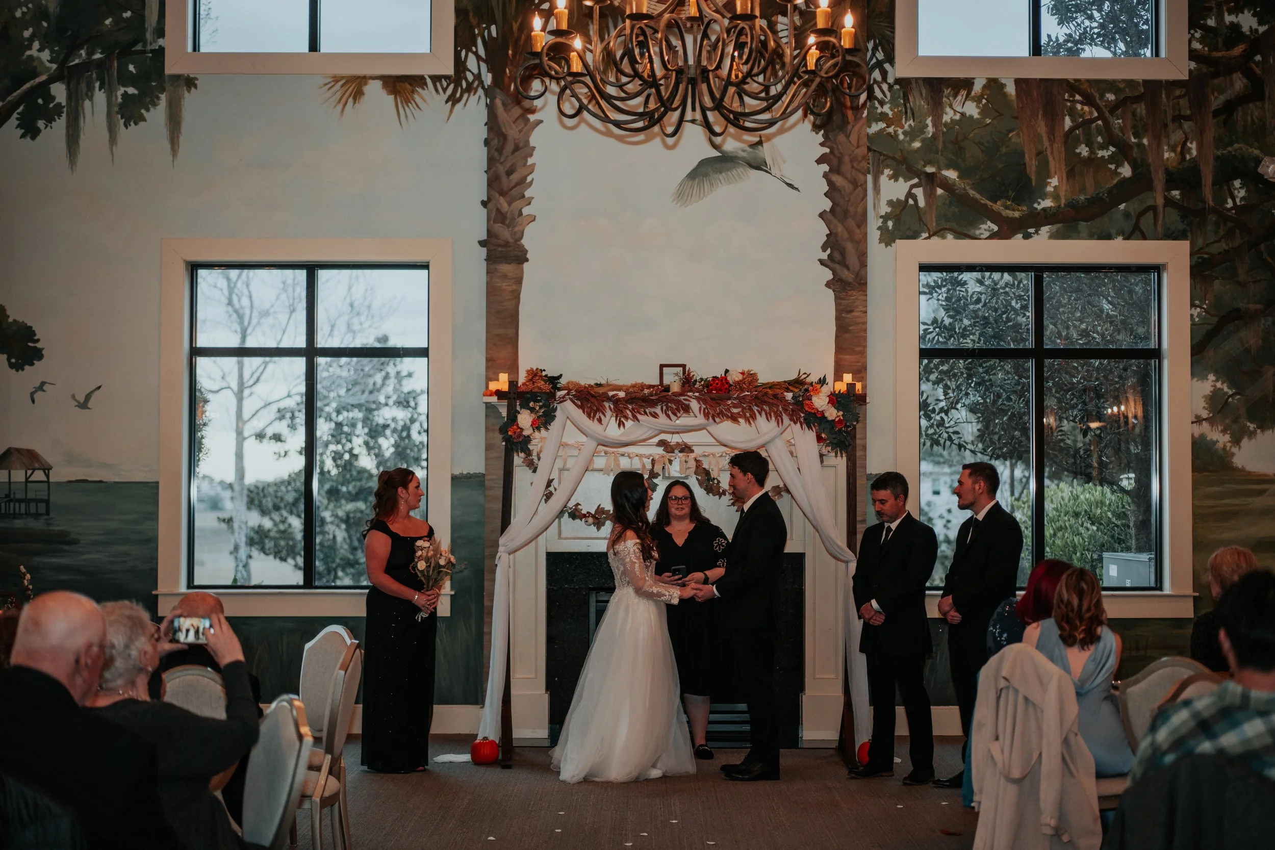 Indoor ceremony with the couple exchanging vows beneath a floral arch framed by painted palm trees and candlelight — planned by Charlotte Wedding Planner.