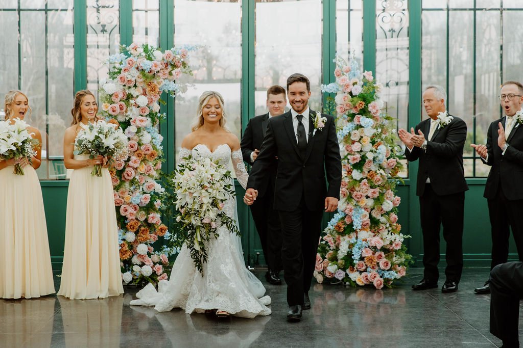 Newlyweds walk hand in hand down the aisle, smiling as guests applaud amid romantic floral arches and soft natural light — planned by Charlotte Wedding Planner at The Little Chapel at Haven Ridge.
