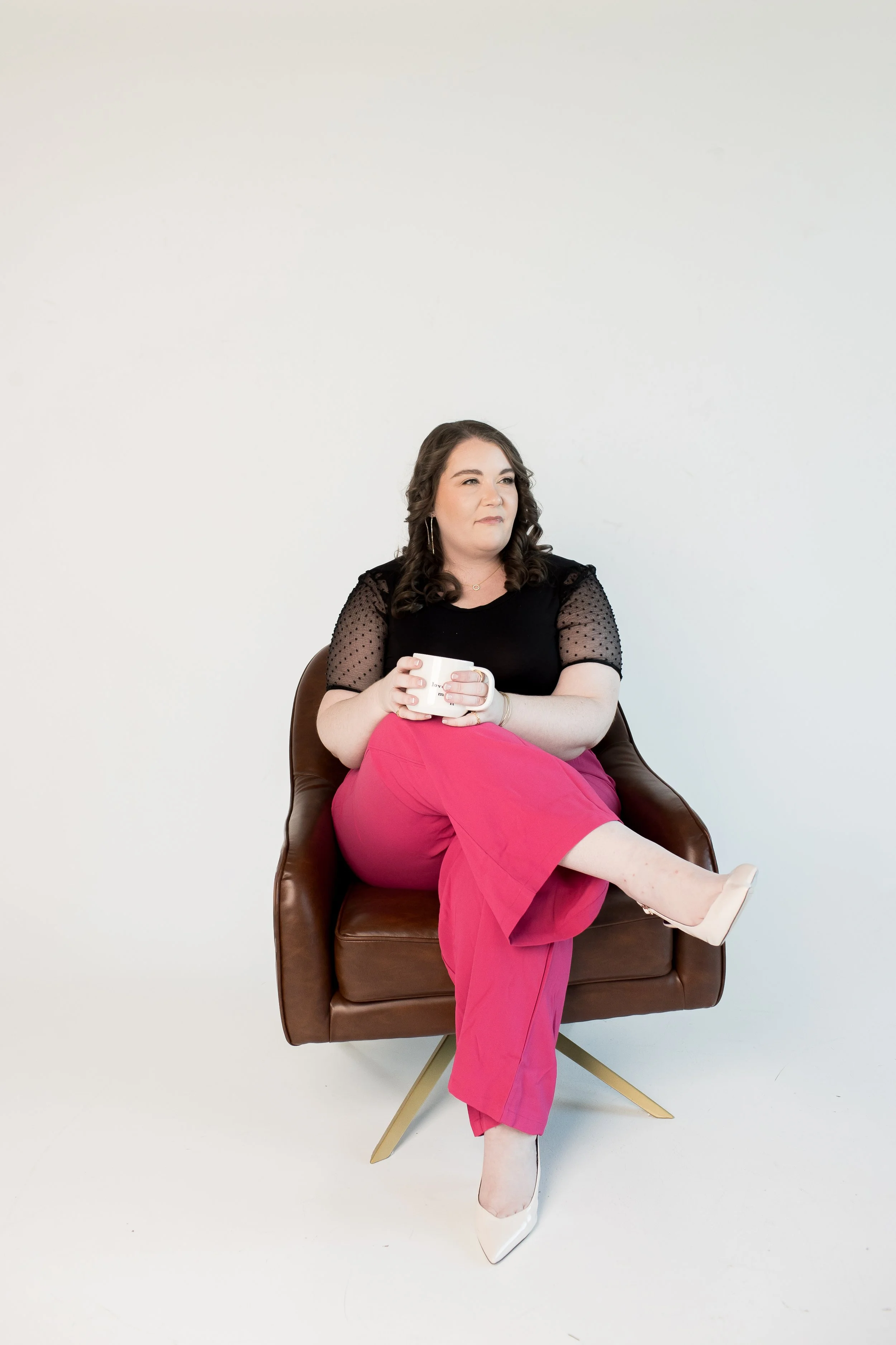 A Charlotte wedding planner sitting in a brown modern chair against a plain white background, holding a white mug, with her legs crossed and wearing a black top with lace sleeves and bright pink pants.