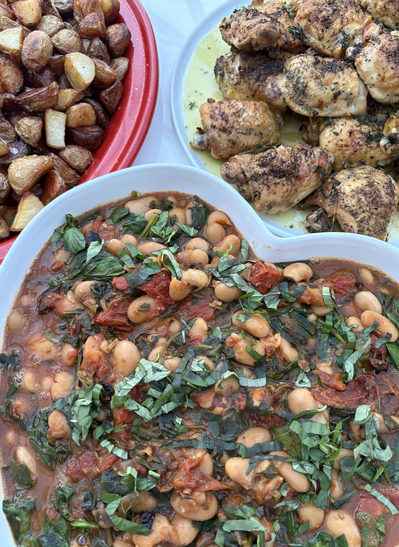 A plate of cooked chicken with herbs, a bowl of chickpea stew topped with chopped herbs, and roasted potatoes.