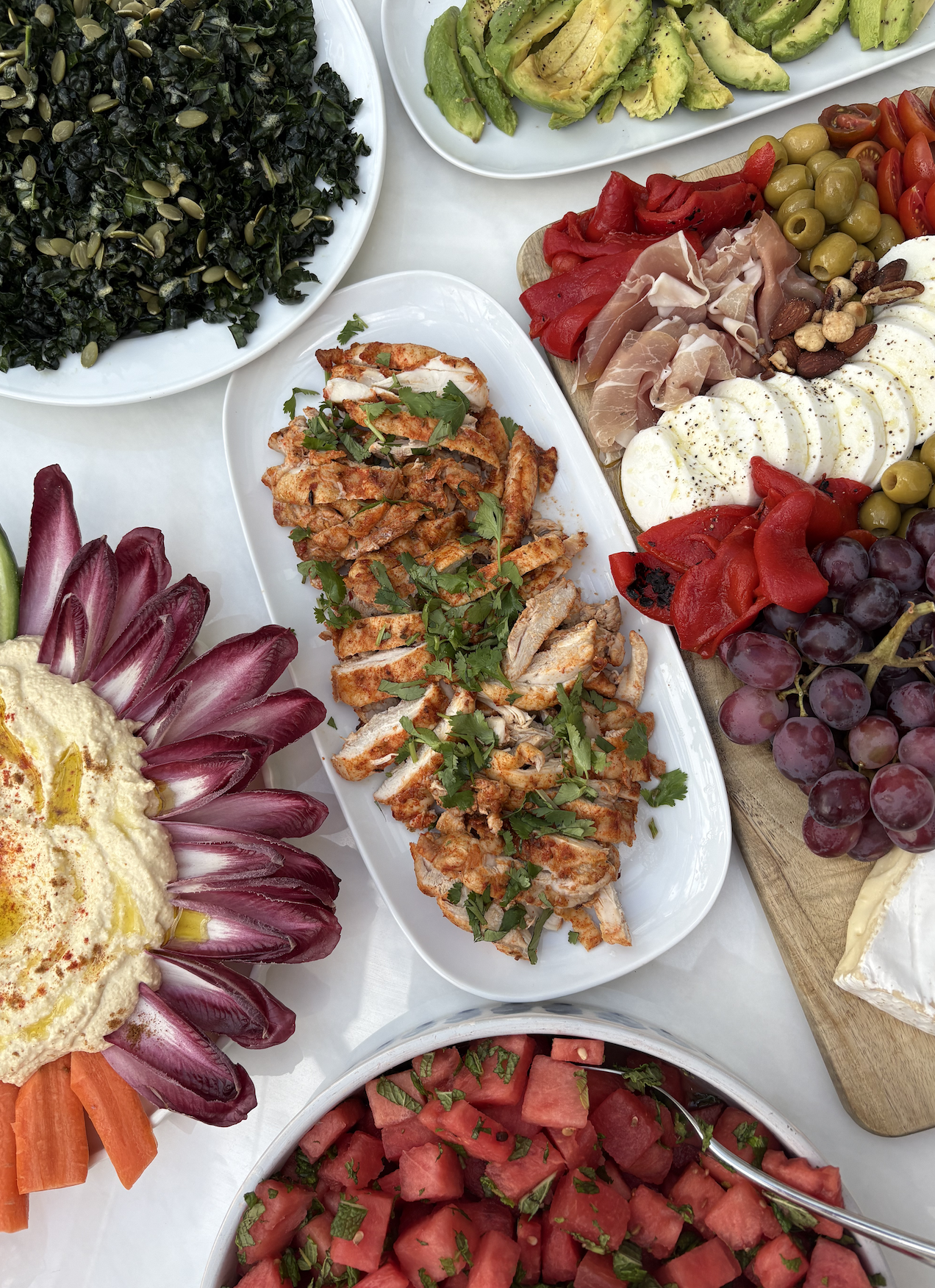 A variety of fresh foods including chopped watermelon, sliced tomatoes, grilled chicken garnished with herbs, sliced avocado, mixed salads with olives, grape clusters, and a charcuterie platter with cheese, meats, nuts, and fruits.