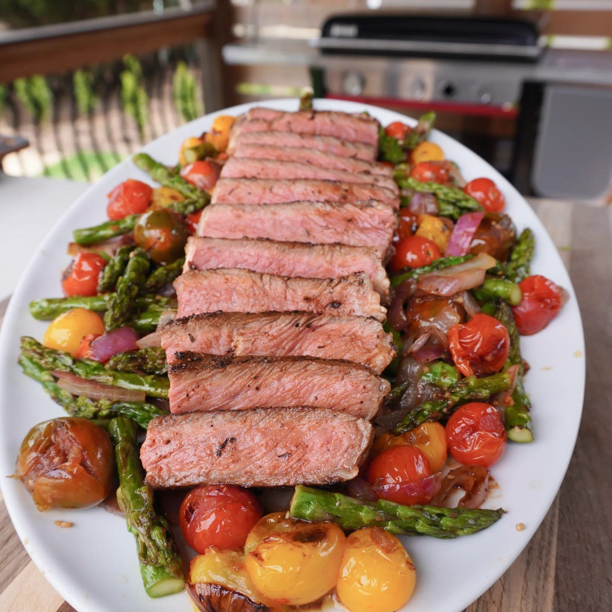 A white plate with sliced cooked steak, cherry tomatoes, yellow heirloom tomatoes, roasted onions, and grilled asparagus on a wooden surface.