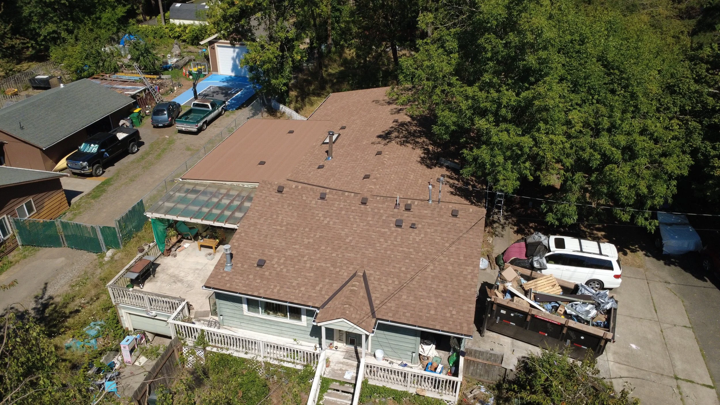 Aerial view of a house with a brown roof, rear deck with items, surrounded by trees and neighboring houses, with parked cars and a large dumpster filled with debris.