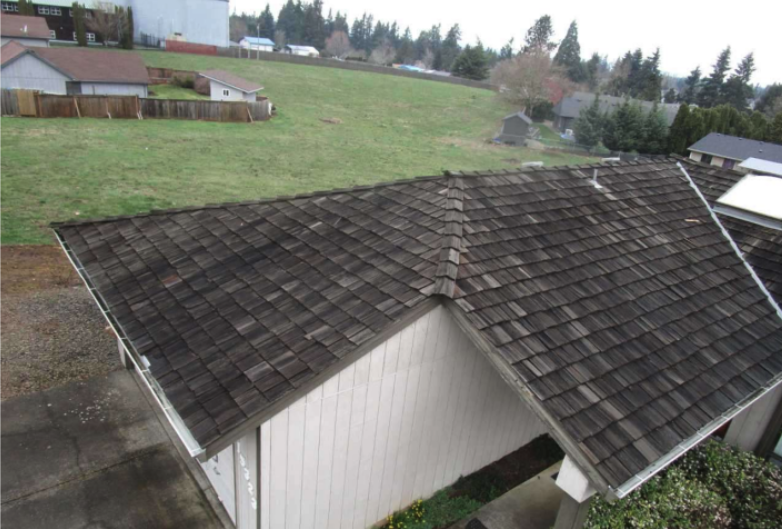 A roof with brown shingles on a residential house, surrounded by other houses and open grassy areas.
