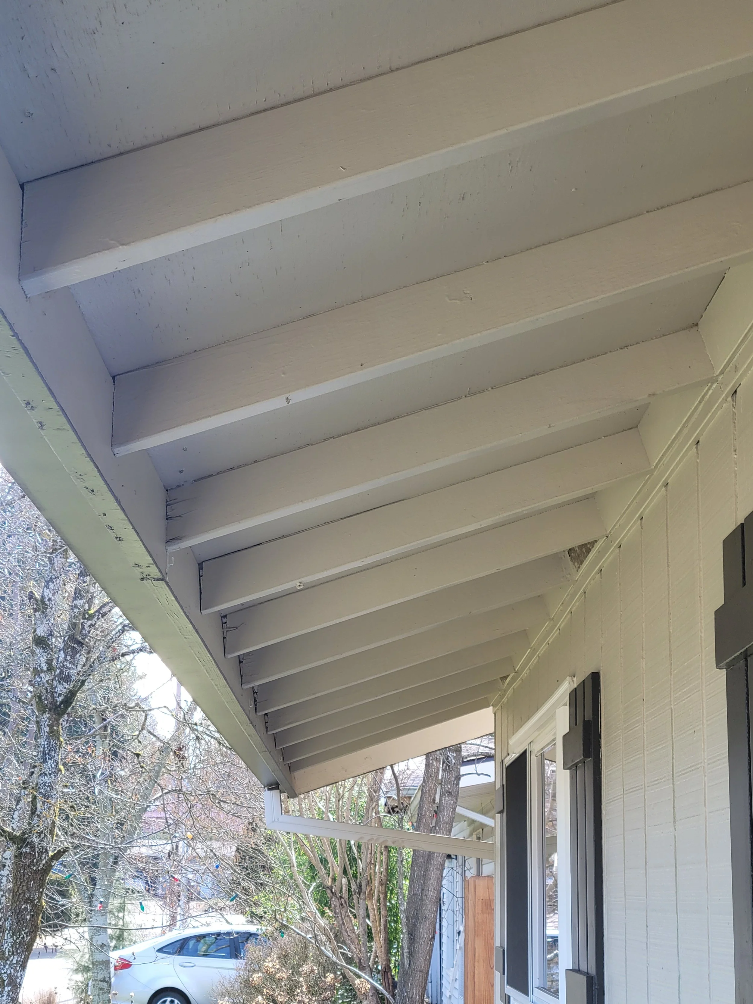 The image shows a close-up view of the porch ceiling of a house, including the wooden beams and the siding of the house. There are some trees and a car visible outside.
