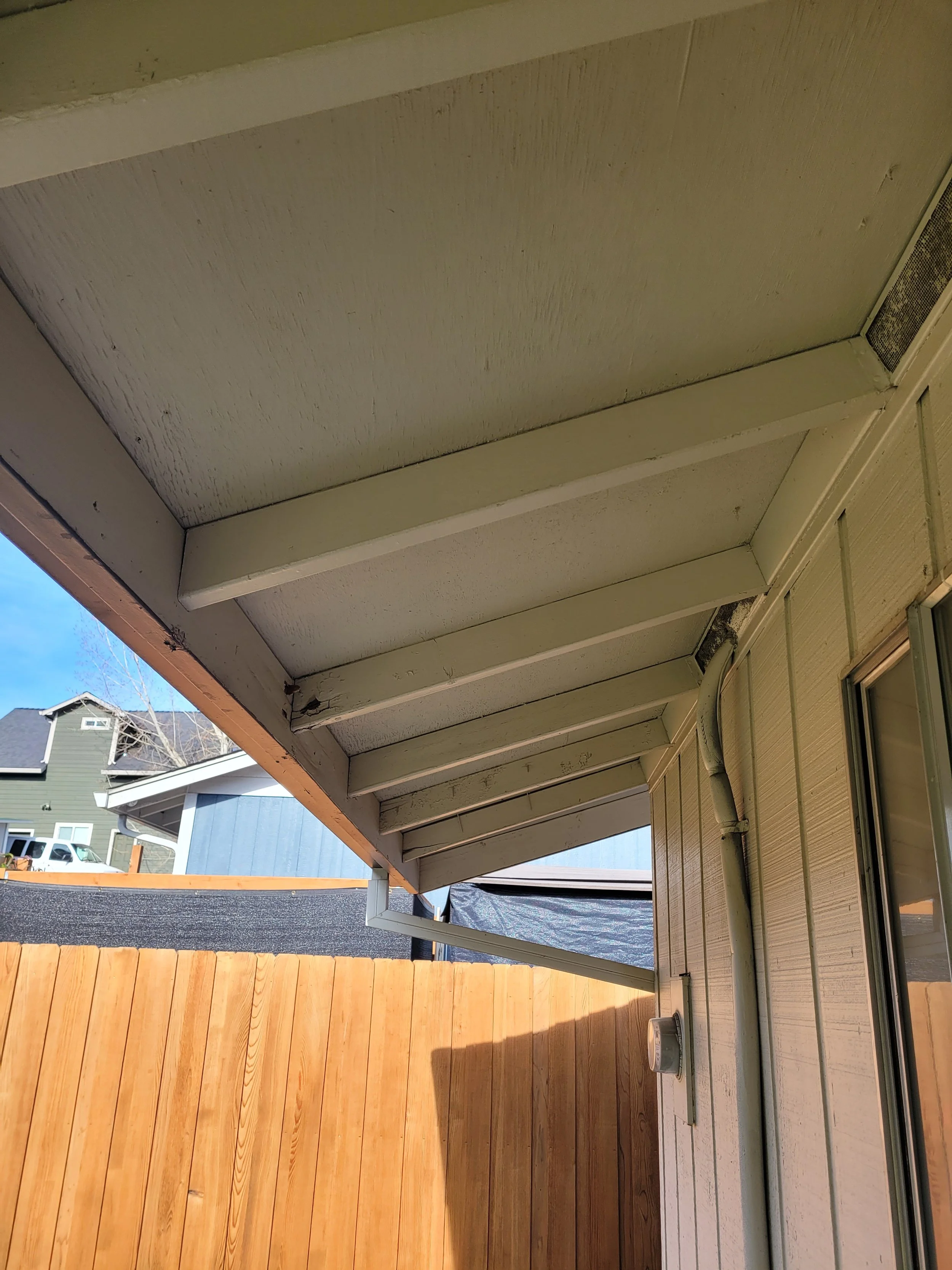 View of the underside of a house's roof overhang featuring exposed beams, with a wooden fence and neighboring houses in the background.