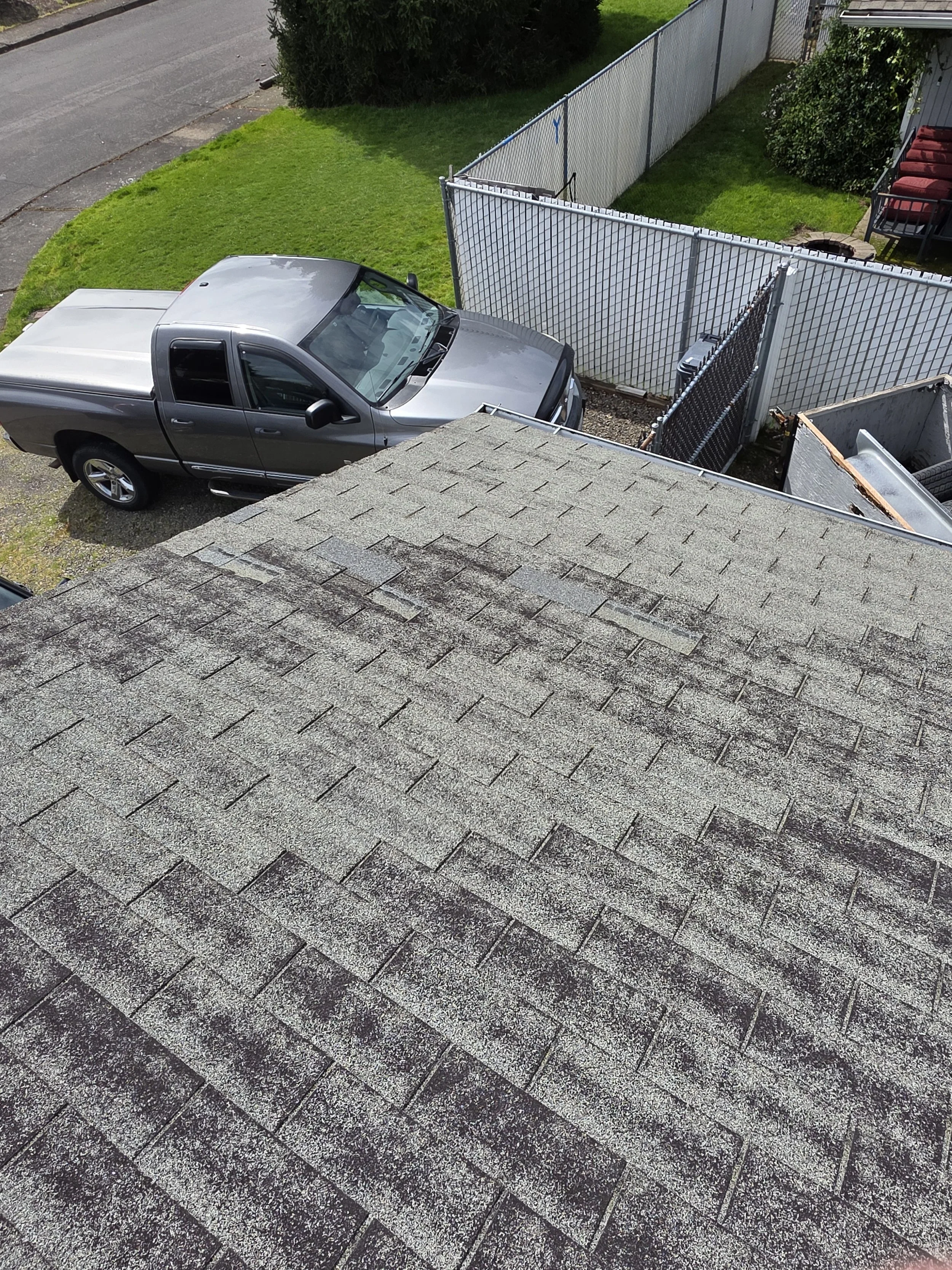 View from above of a gray pickup truck parked on a gravel driveway behind a house with shingled roof, white fence, green grass, and neighboring houses.