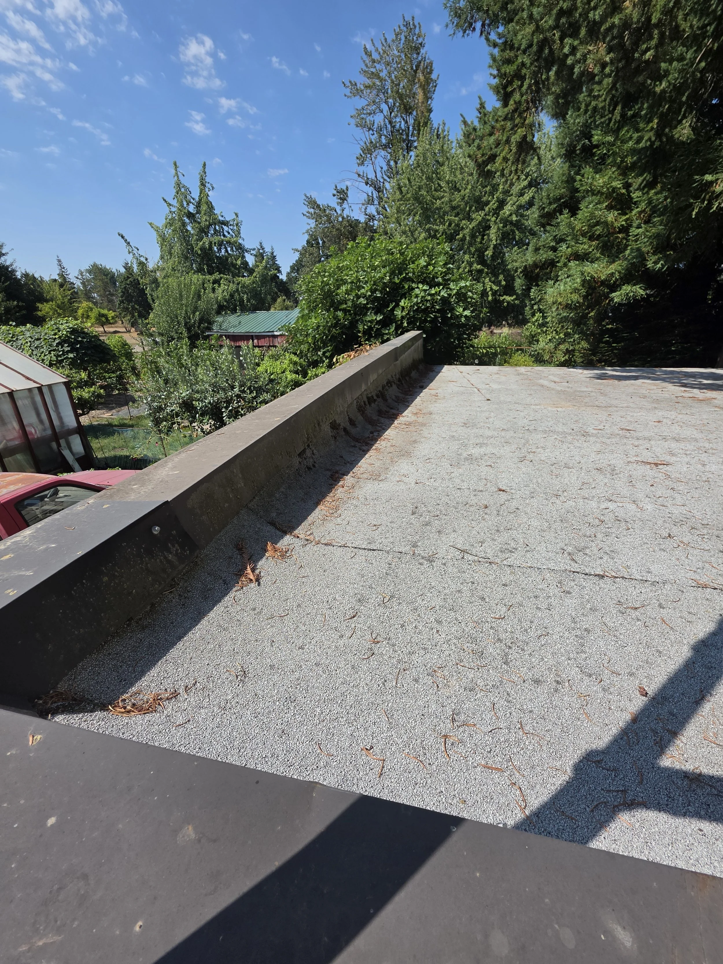 View of a flat, gravel rooftop with a concrete curb, surrounded by green trees and a partly cloudy blue sky.