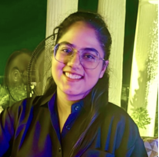 A young woman with glasses smiling at the camera. She has dark hair tied back and is wearing a dark shirt. The background includes a standing fan, white pillars, and a window with sunlight.