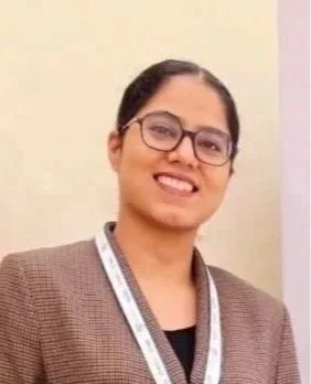 A young woman with glasses smiling at the camera. She has dark hair tied back and is wearing a dark shirt. The background includes a standing fan, white pillars, and a window with sunlight.