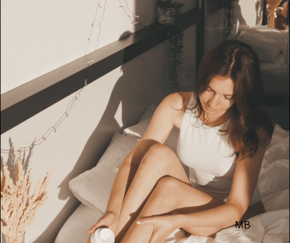 A woman sitting on a bed applying lotion to her leg, in a sunlit room with plants and a light-colored wall.
