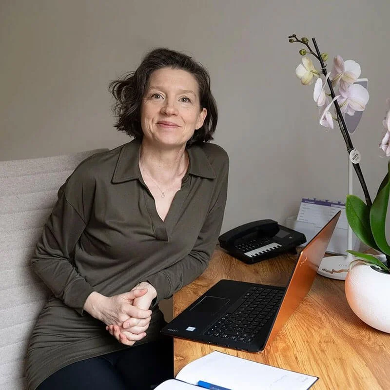 Porträt von Sandra Stark. Sie schaut freundlich, lachend direkt in die Kamera, sitzt am Schreibtisch vor Laptop, trägt eine dunkelgrün/ braune Bluse, hat braune, kinnlange Haare.