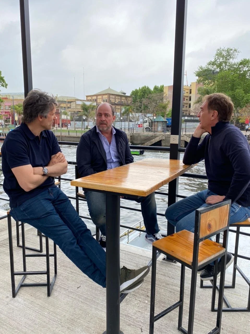 Tres hombres sentados en una mesa de madera en un barco, conversando, con un río y edificios de fondo.