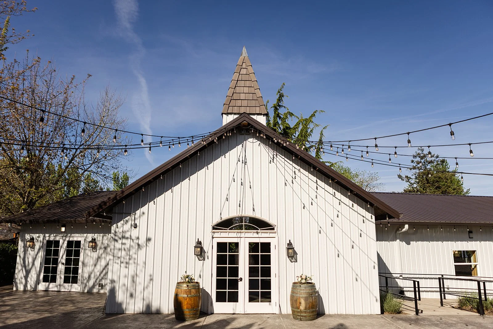 Photo of a white barn with a peaked roof and a small tower, decorated with string lights, with two barrels and flowers in front, against a clear blue sky.