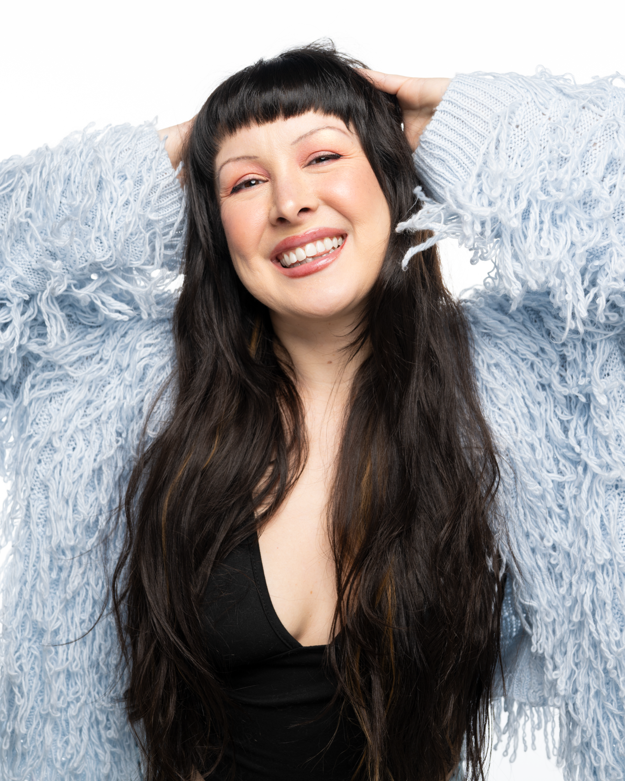 A woman with long dark hair and a short fringe, smiling with teeth visible, wearing a light-colored textured sweater, against a plain white background.