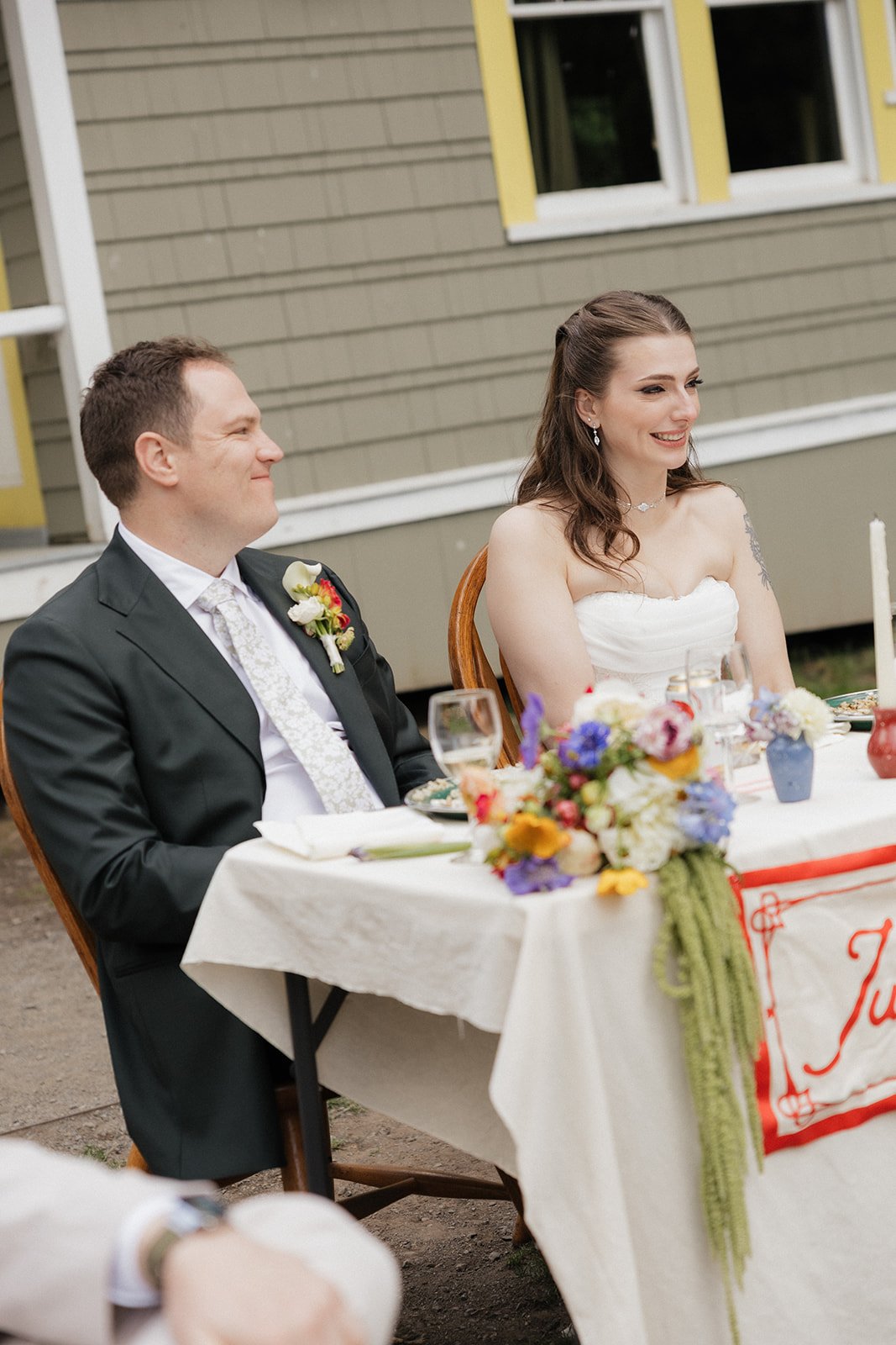 A bride and groom sitting at a decorated wedding reception table outdoors, smiling. The groom is in a black suit with a white tie and boutonniere, and the bride is in a strapless white dress with jewelry. The table has a floral centerpiece, wine glas