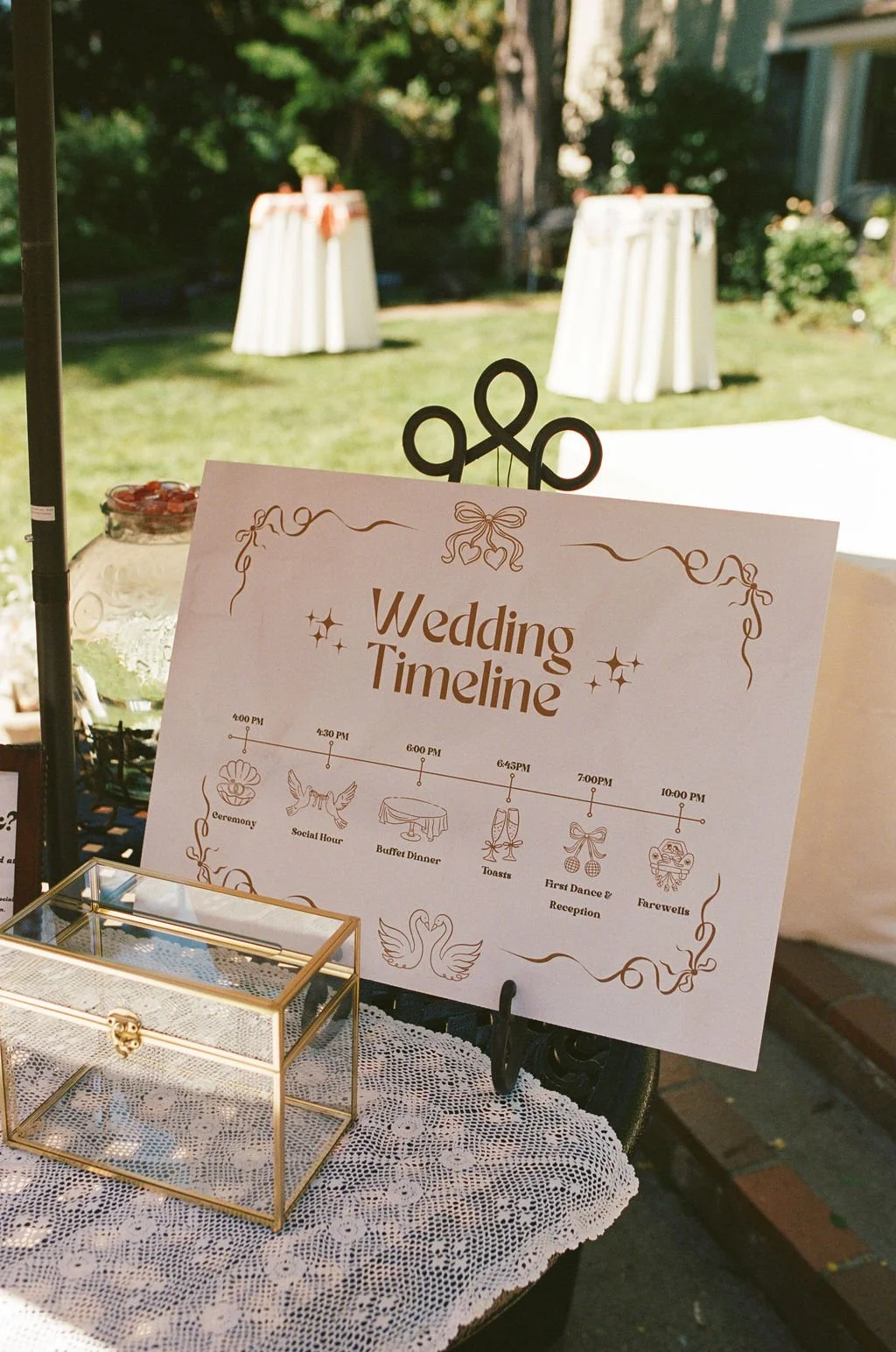 Wedding timeline sign displaying schedule of events including ceremony at 4:00 PM, social hour at 4:30 PM, buffet dinner at 6:00 PM, toasts at 6:45 PM, first dance and reception at 7:00 PM, and farewells at 10:00 PM, on a table with a lace cloth and 