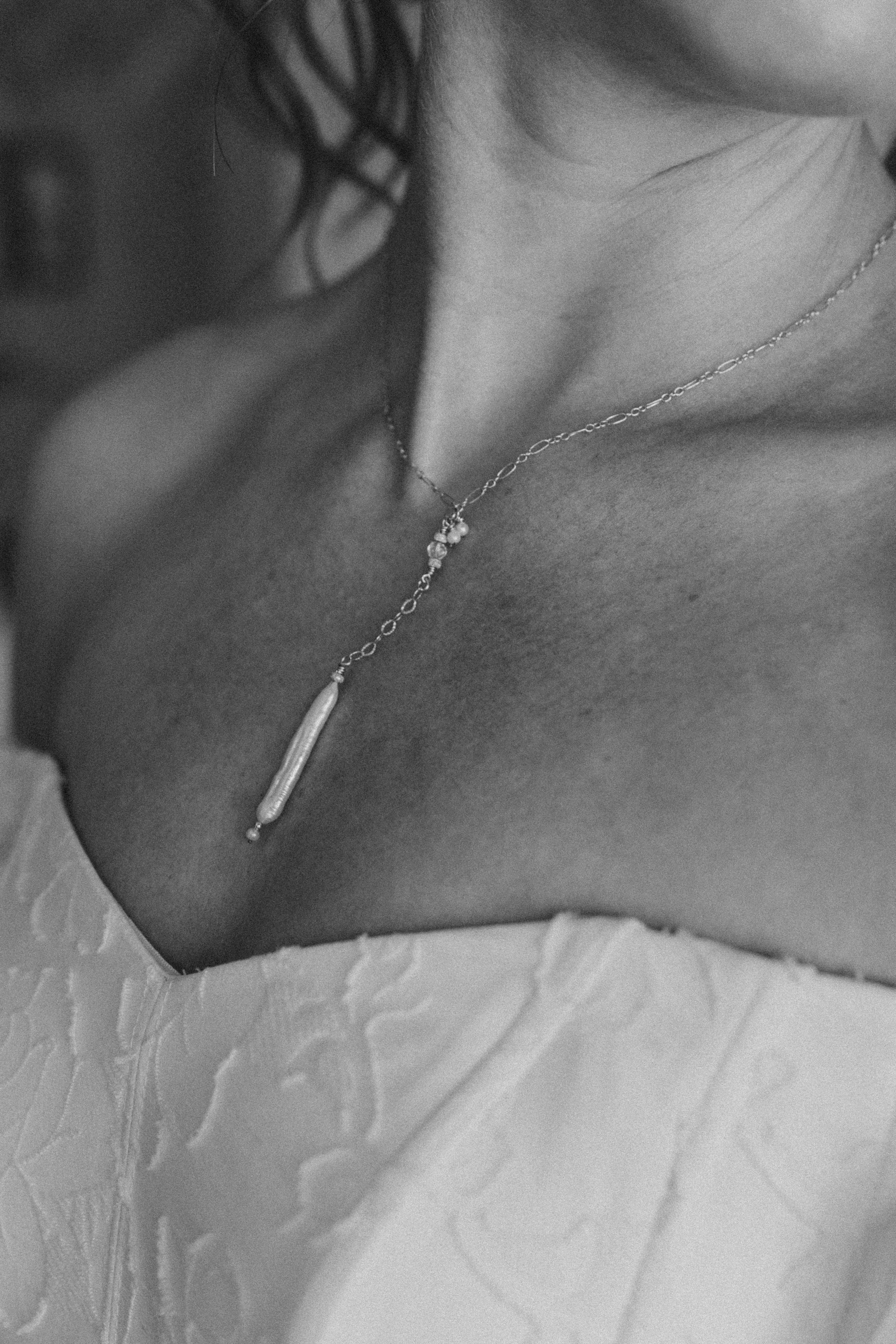 Close-up black and white photograph of a woman's neck and collarbone with a delicate chain necklace and textured fabric in the background.