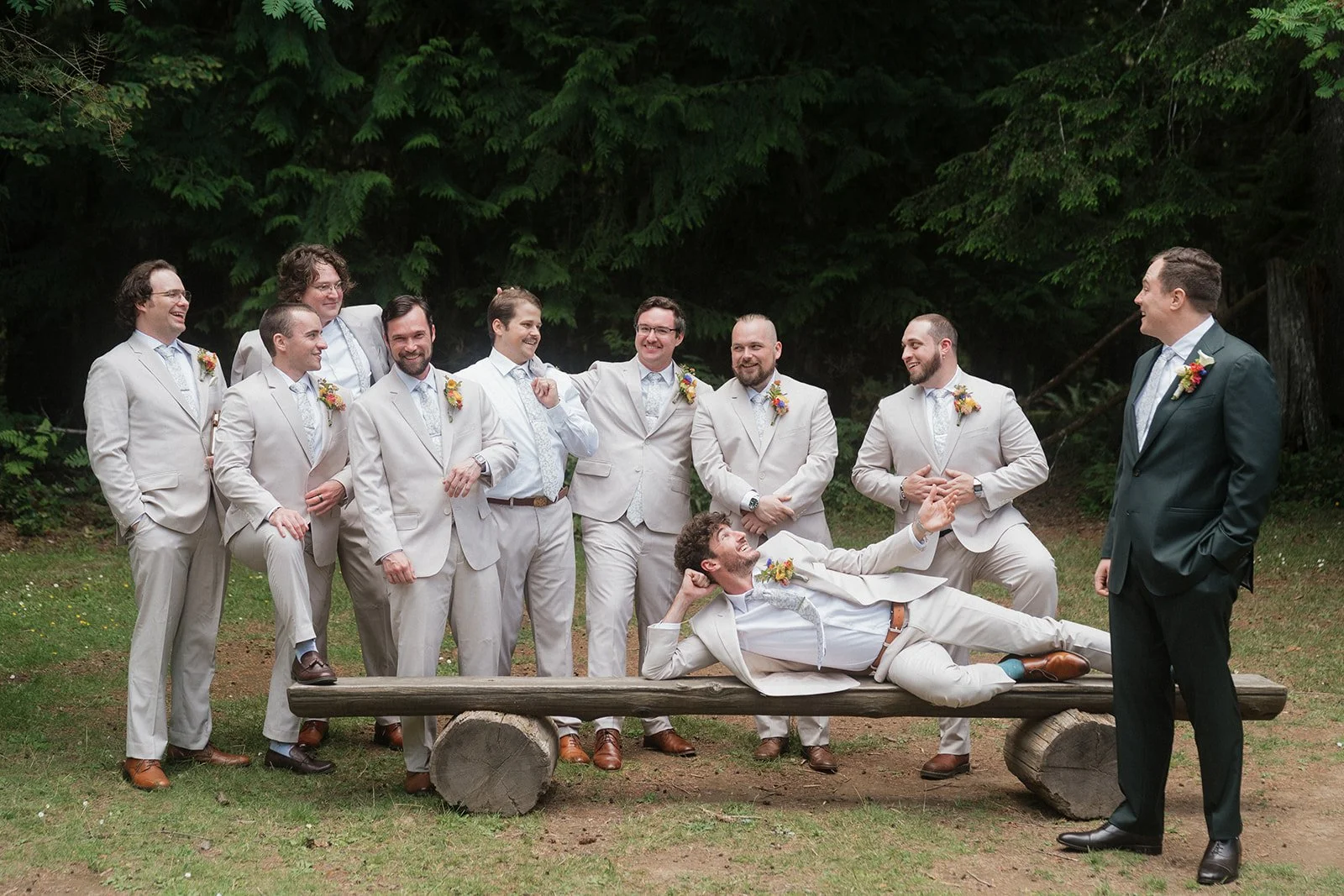 A group of men in light-colored suits and one man in a dark suit are posing outdoors in front of green trees. They are smiling and laughing, with one man lying on a wooden bench in front of the group, striking a playful pose.
