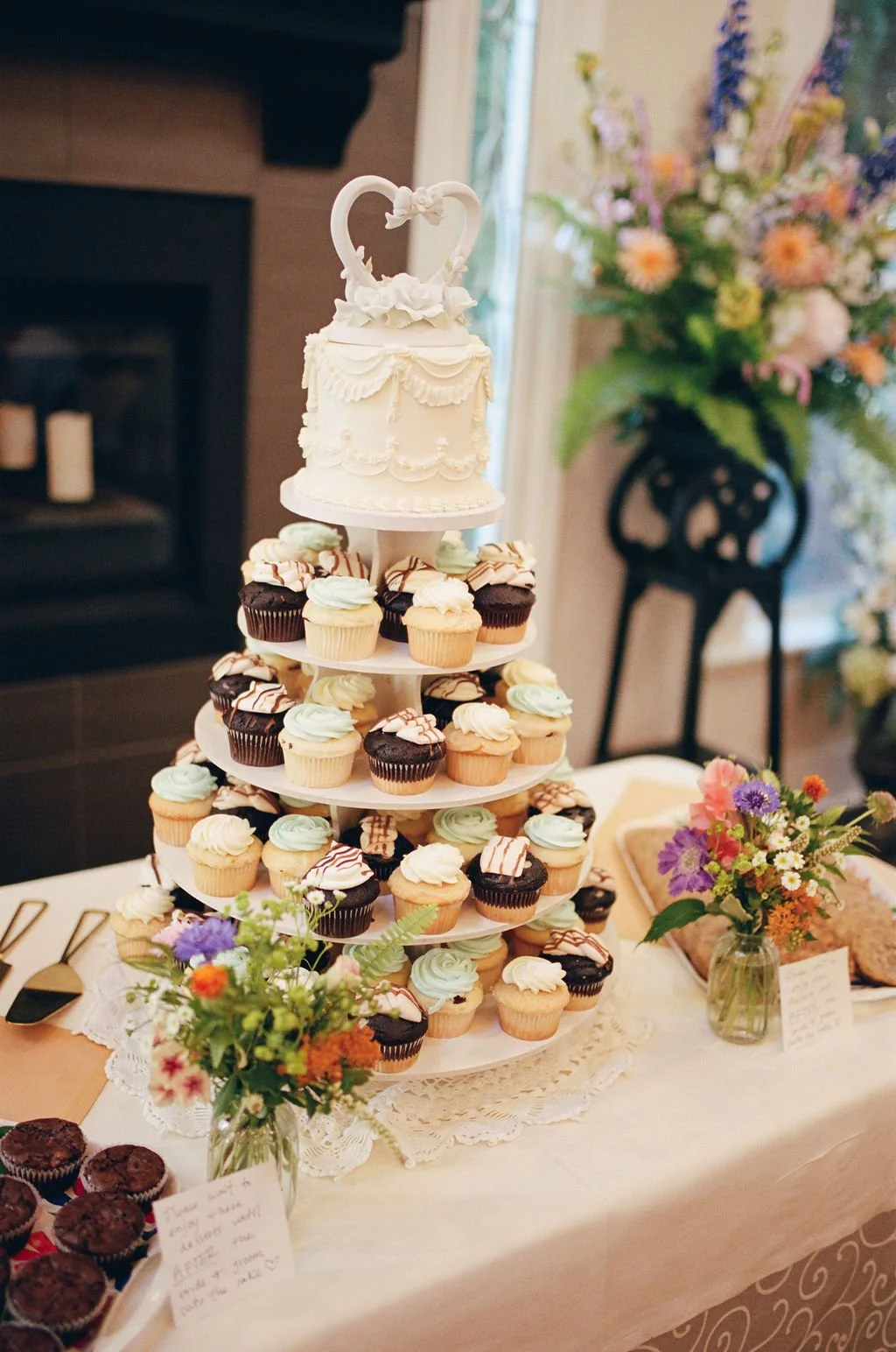 A wedding cake with a swan topper, placed on a multi-tiered stand surrounded by cupcakes with pastel-colored frosting. The table has flowers and a lace doily.