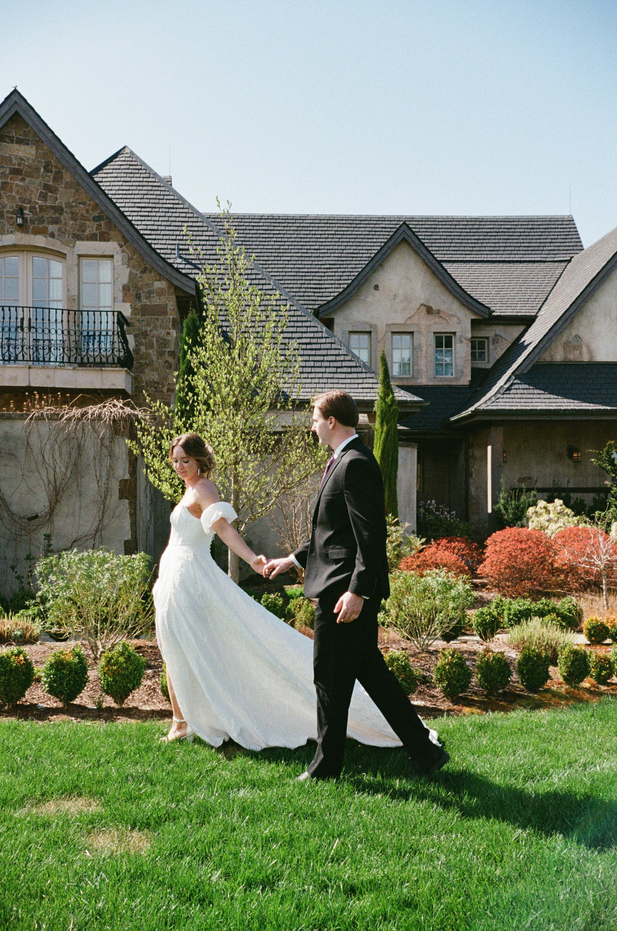 A bride and groom walking hand in hand in a garden with lush green grass, bushes, and flowering plants, in front of a large house with stone and stucco exterior and multiple gabled roofs.