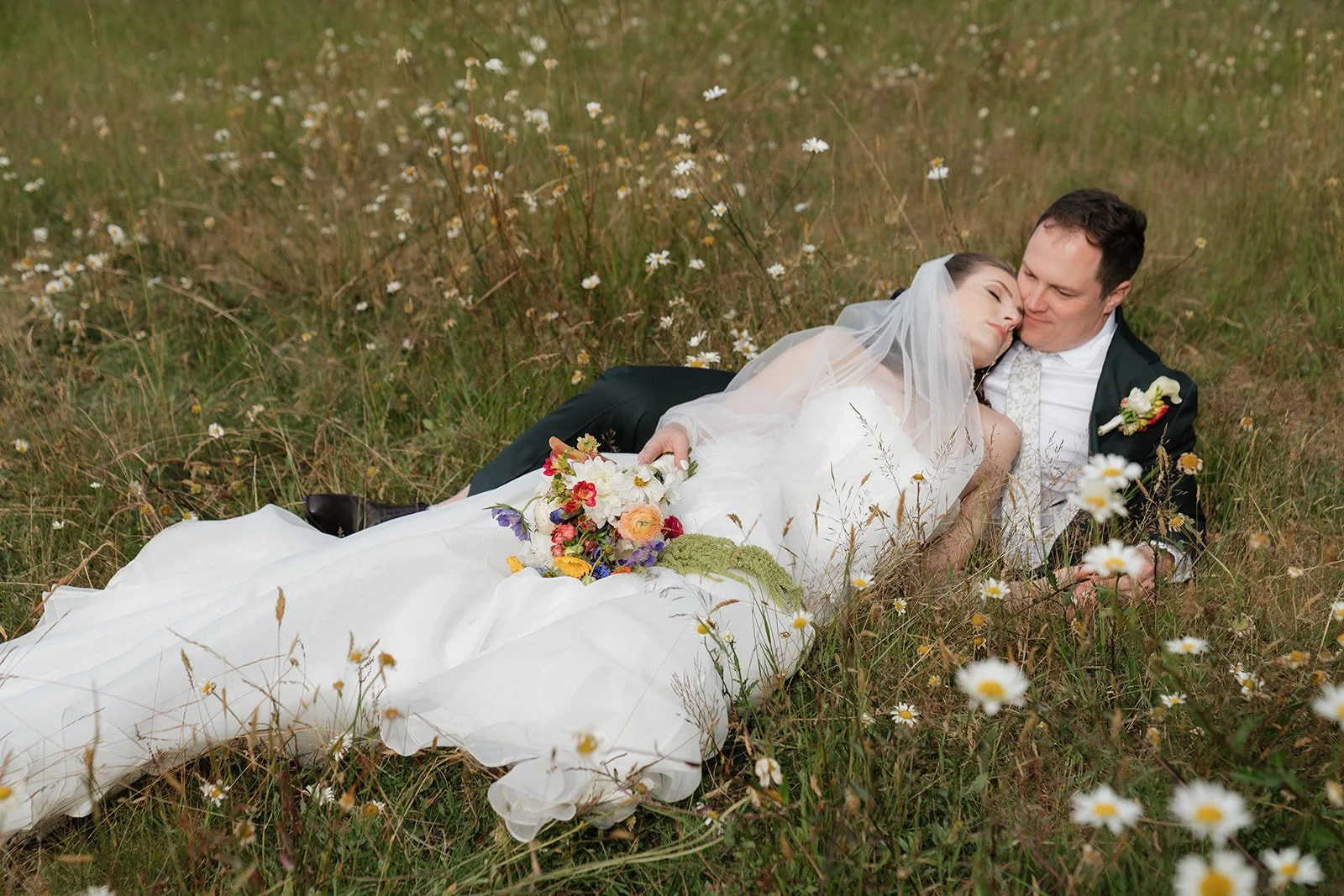 A bride and groom lying together on a grassy field with daisies, embracing and kissing. The bride is holding a bouquet of colorful flowers, and she wears a white wedding dress with a veil. The groom is dressed in a dark suit with a white shirt and bo