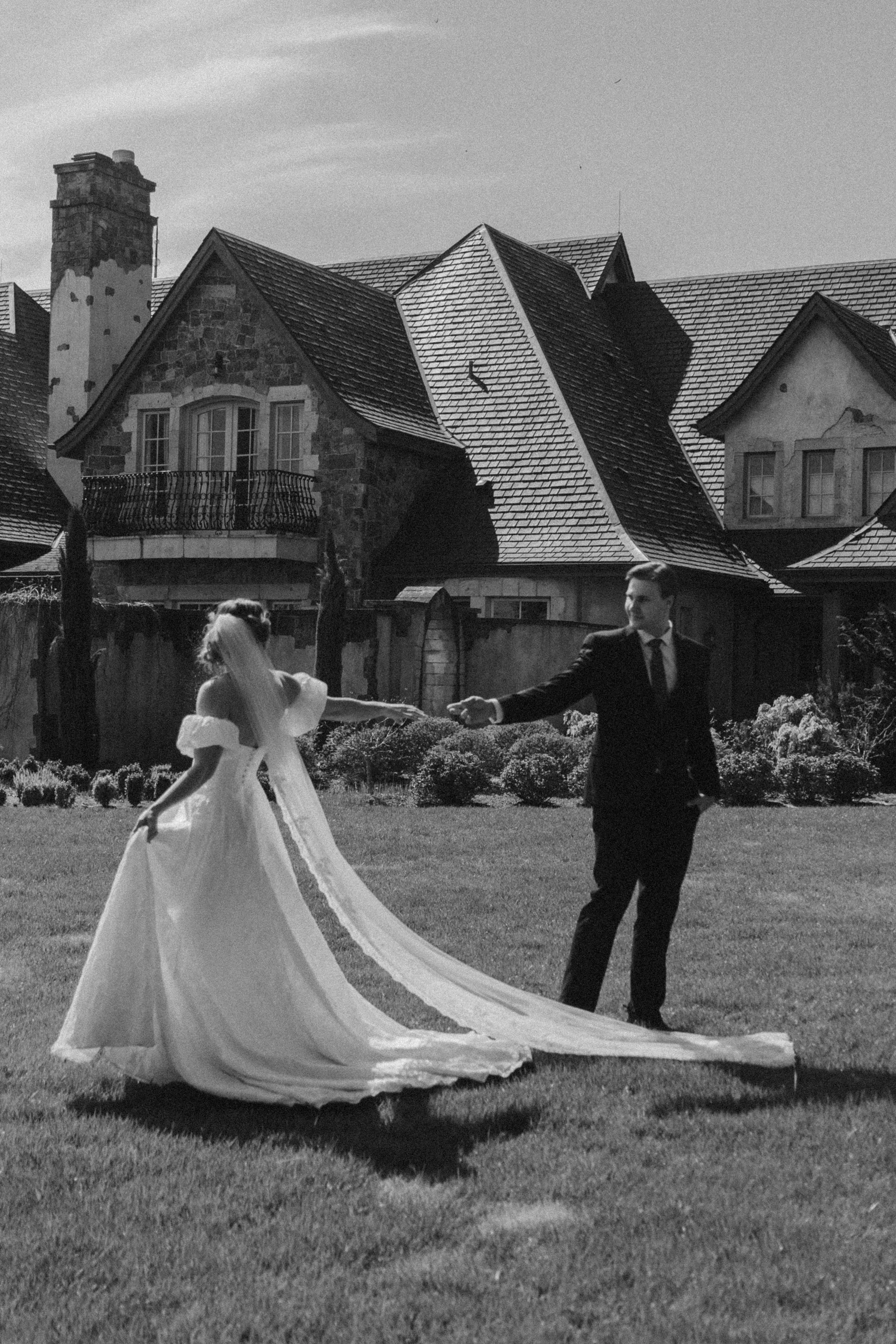 A bride and groom dancing outside in front of a house with multiple gabled roofs, a brick chimney, and stone walls in a black and white photograph.