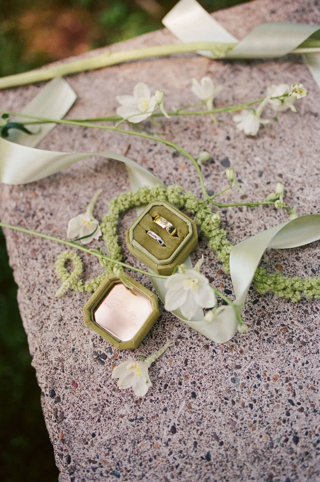 A ring in a green velvet box placed on a gray stone surface, surrounded by white flowers and green leaves.