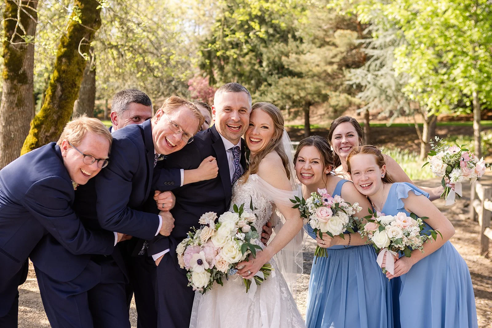 A group of people at a wedding, including the bride and groom, standing outdoors among trees, all smiling and holding bouquets of flowers.