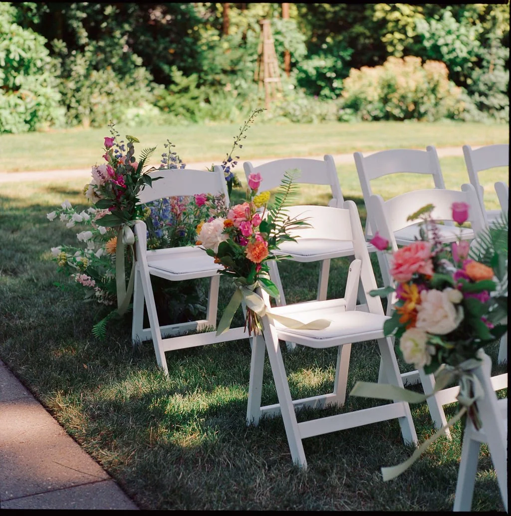 White folding chairs decorated with bouquets of colorful flowers, arranged outdoors in a garden setting for a wedding or event.