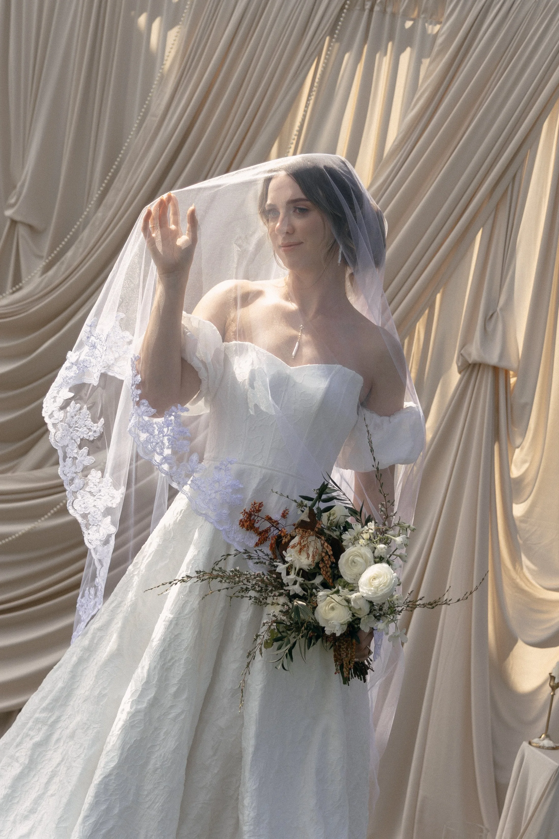 A bride in a white wedding dress holding a bouquet of white roses, standing in front of beige draped curtains, with a sheer veil over her face and dark hair styled in loose waves.