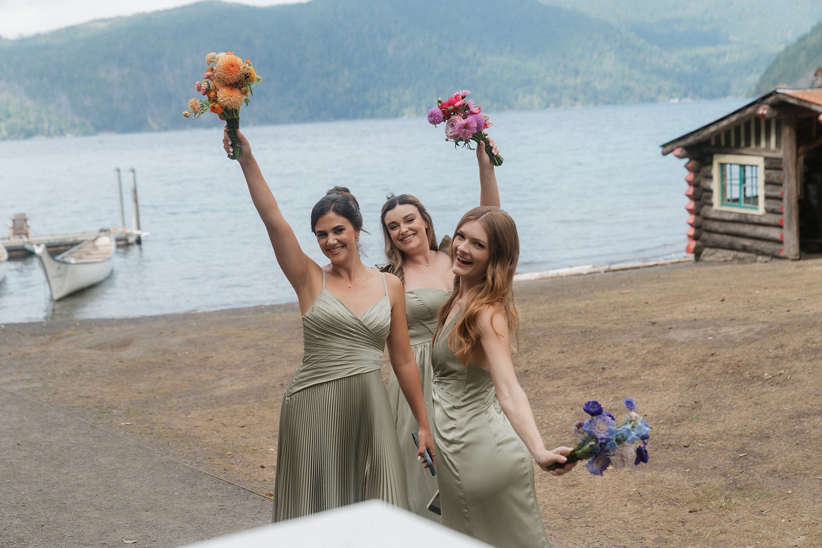 Three women in matching beige dresses celebrating outdoors near a lake with boats and mountains, holding colorful bouquet flowers.