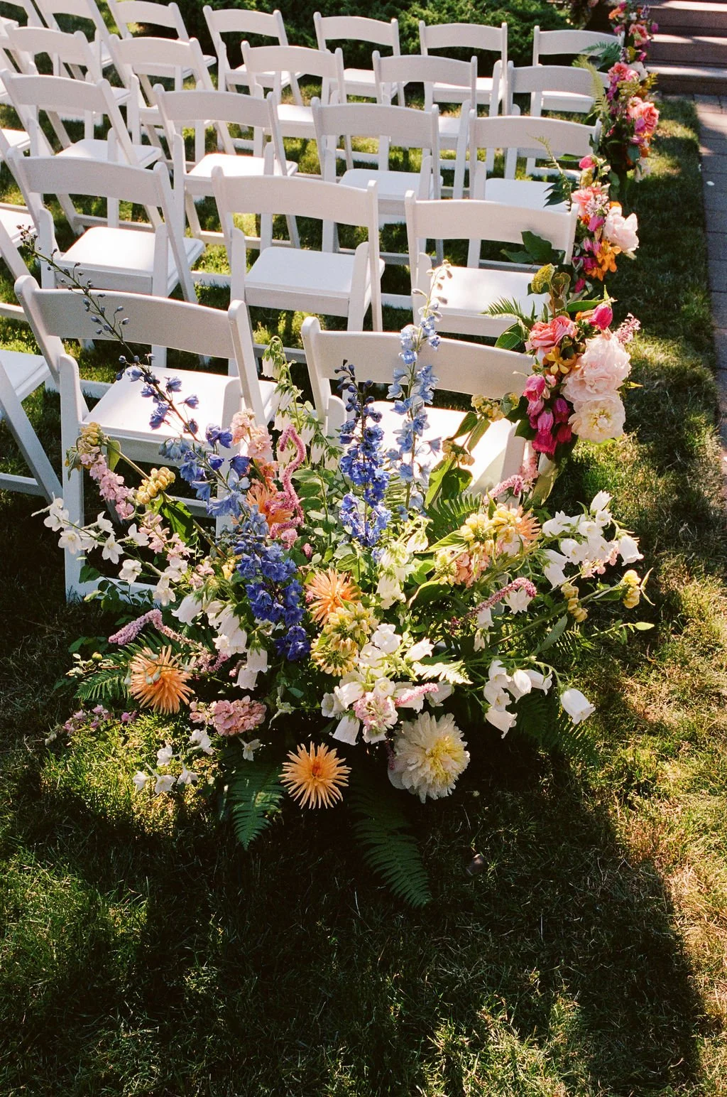 White folding chairs arranged outdoors with a floral arch made of pink, white, and purple flowers, set on a grassy area for a wedding ceremony.