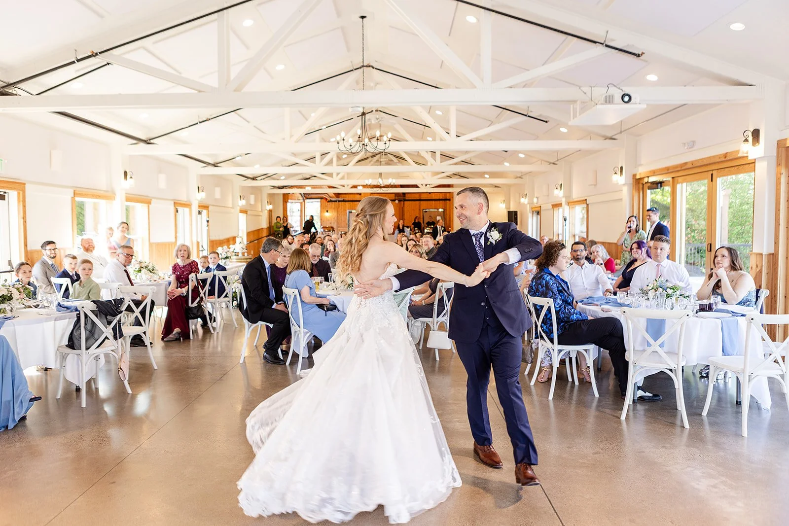 Bride and groom dancing at a wedding reception, with guests seated at decorated tables in a bright, spacious hall.