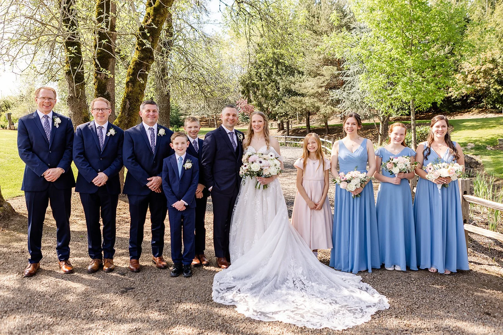 A wedding party outdoors under trees, including the bride and groom, groomsmen in navy suits, bridesmaids in blue dresses, and children. The bride holds a bouquet, and all are smiling.