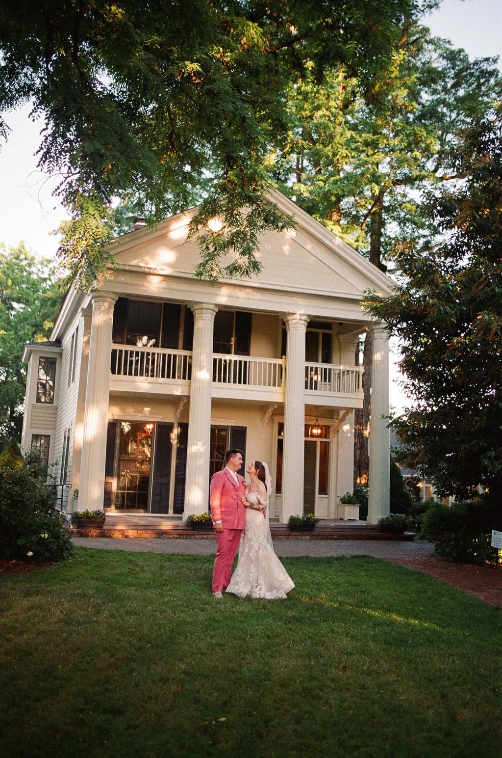 A bride and groom stand on a lawn in front of a large white house with tall columns, surrounded by trees, during sunset.
