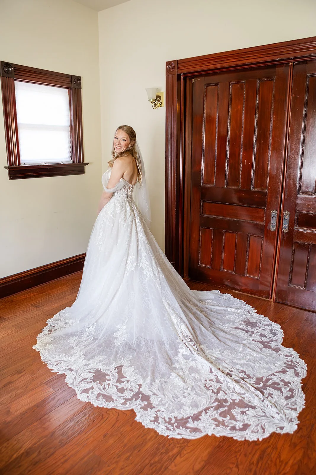 A smiling woman in a wedding dress standing in a room with wooden trim and floor, next to a window and double wooden doors.