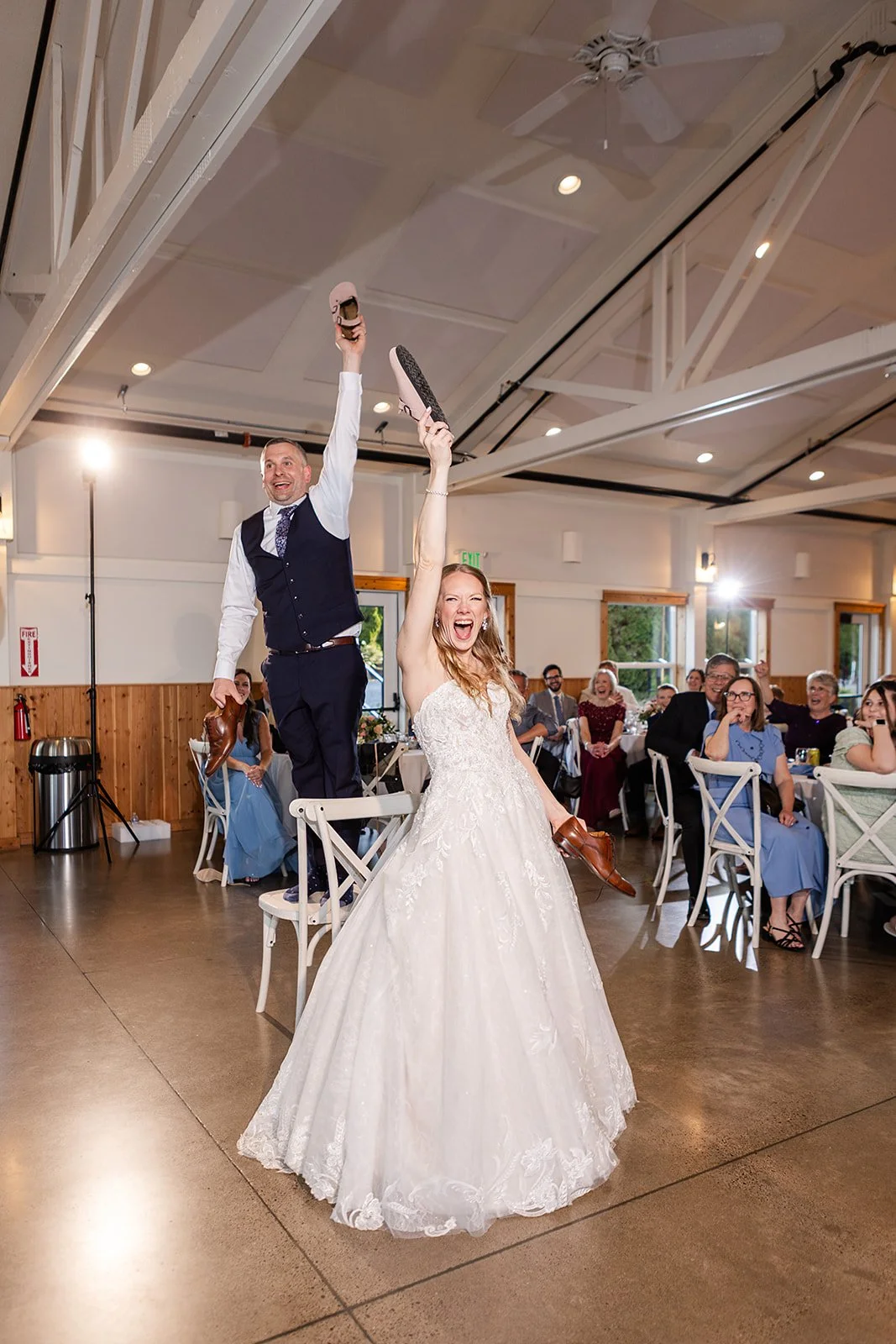 Wedding reception with a bride and groom holding shoes while standing on chairs, guests seated at tables, smiling and cheering in a rustic venue.