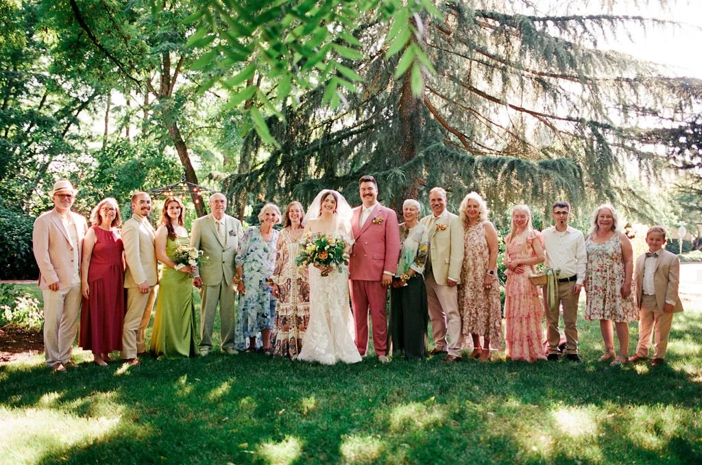 A wedding party outdoors with the bride and groom in the center, surrounded by family and friends on a green lawn, with trees and sunlight in the background.