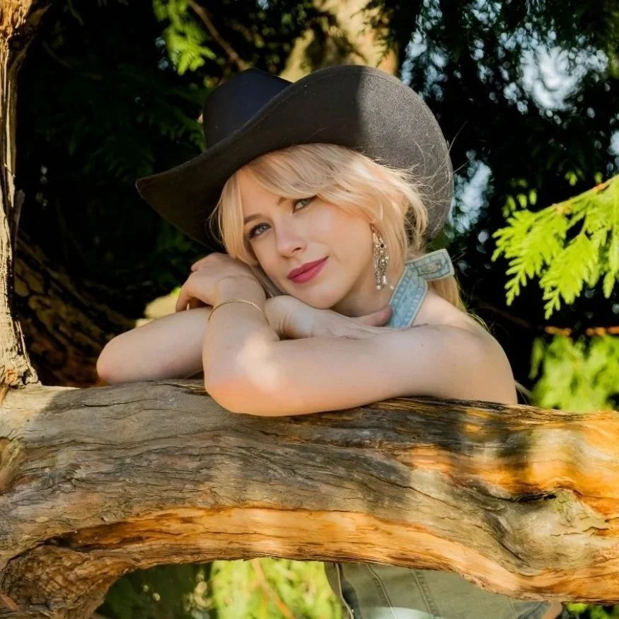 A woman with blonde hair, wearing a large black hat, earrings, and a sleeveless top, rests her head on her arms on a tree branch outdoors, surrounded by green leaves.