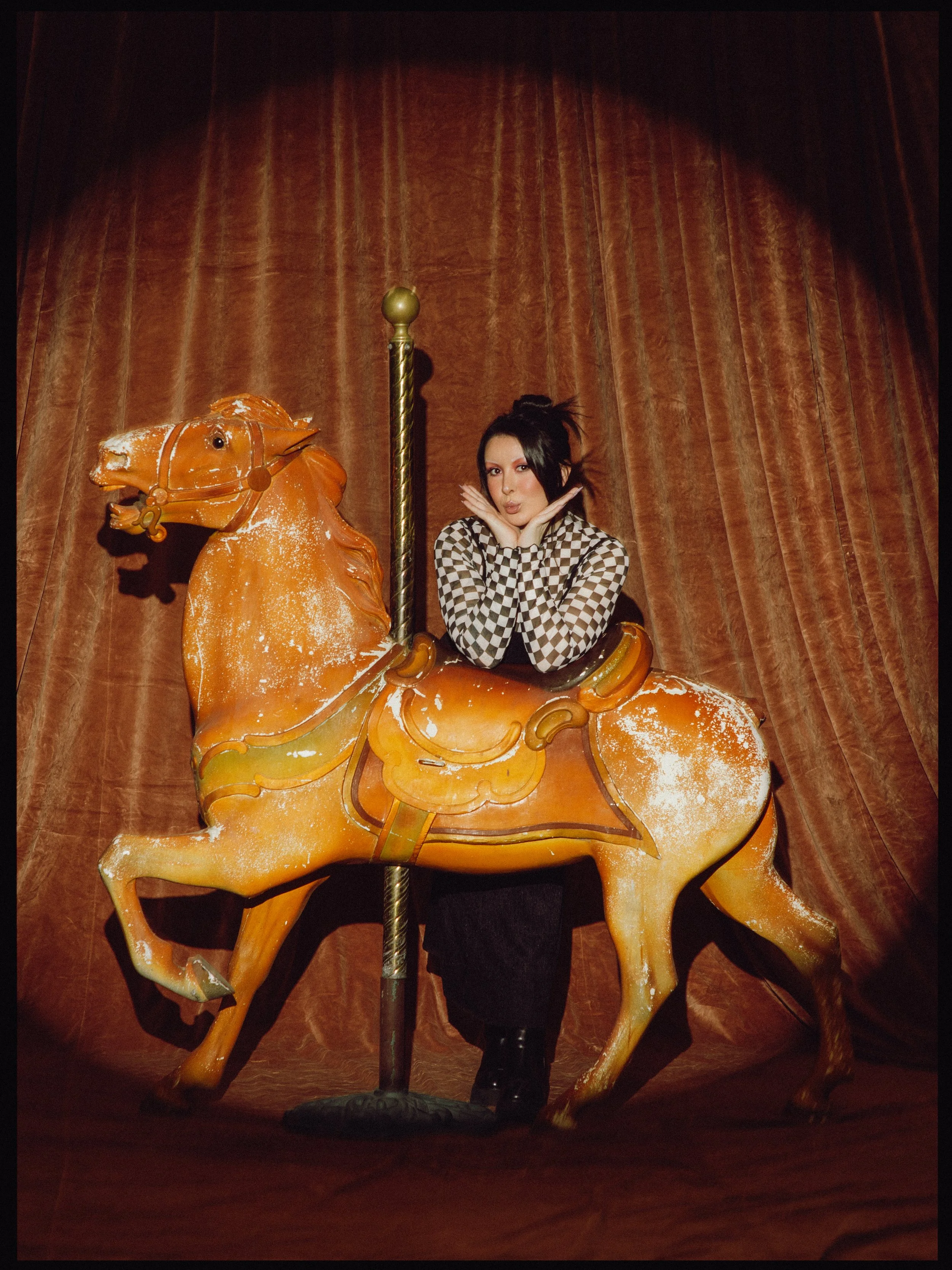 An amusement park carousel horse with a person sitting on it, within a circular, vintage-style setting with brown curtains in the background.