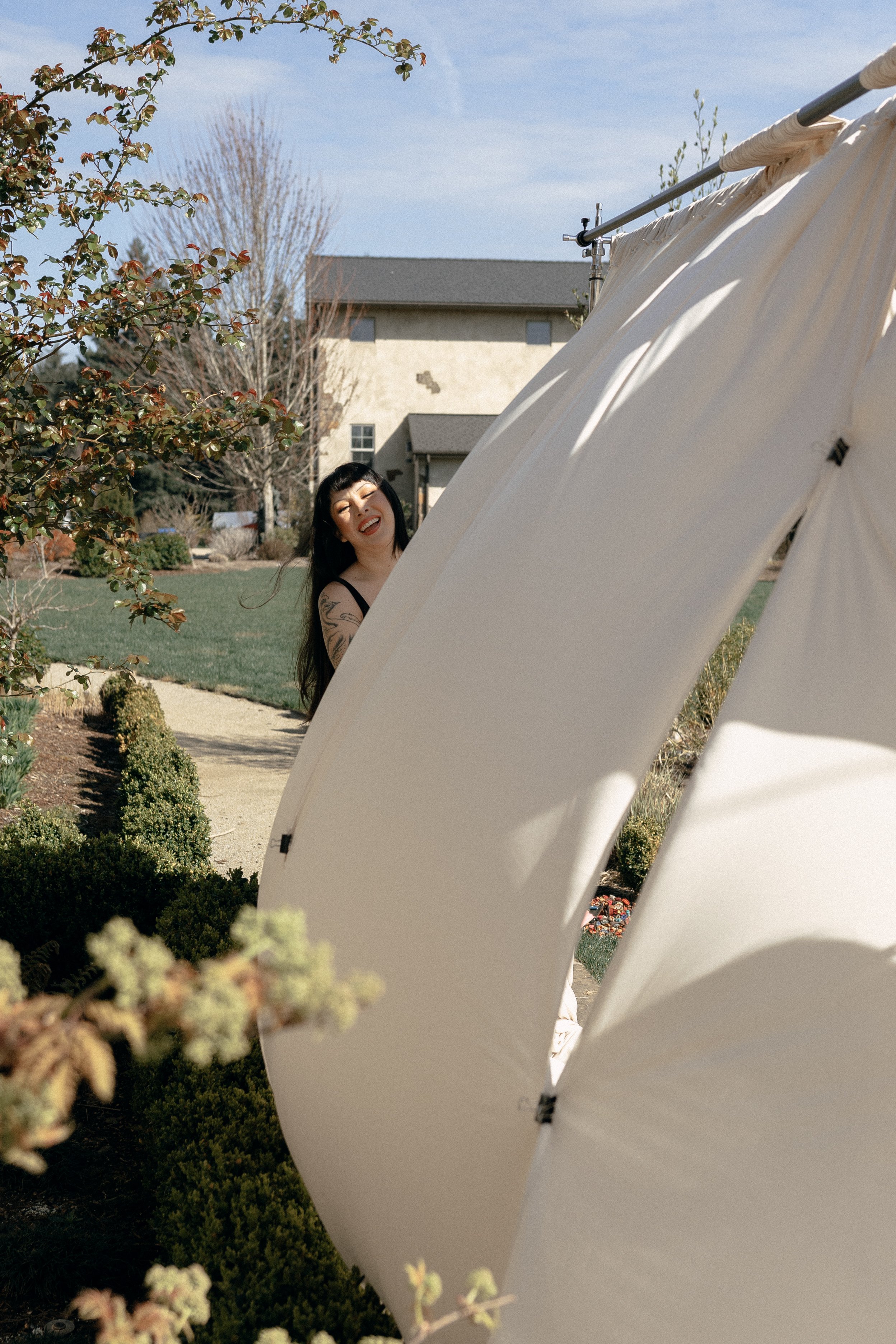 A woman with long dark hair and tattoos on her arm, smiling and looking at the camera, standing outdoors near white tents in a garden with bushes and trees, with a house in the background under a partly cloudy sky.
