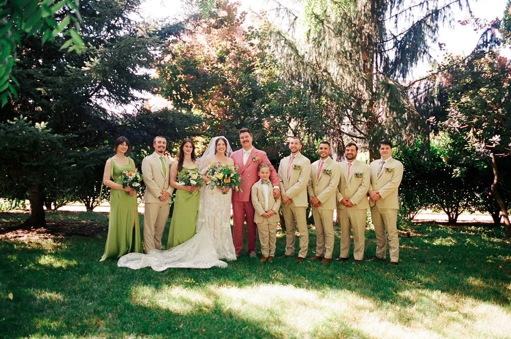 A wedding party outdoors with a bride in a white gown with a veil, a groom in a pink suit, and seven bridesmaids and groomsmen in beige suits and green dresses, standing on grass among tall trees.