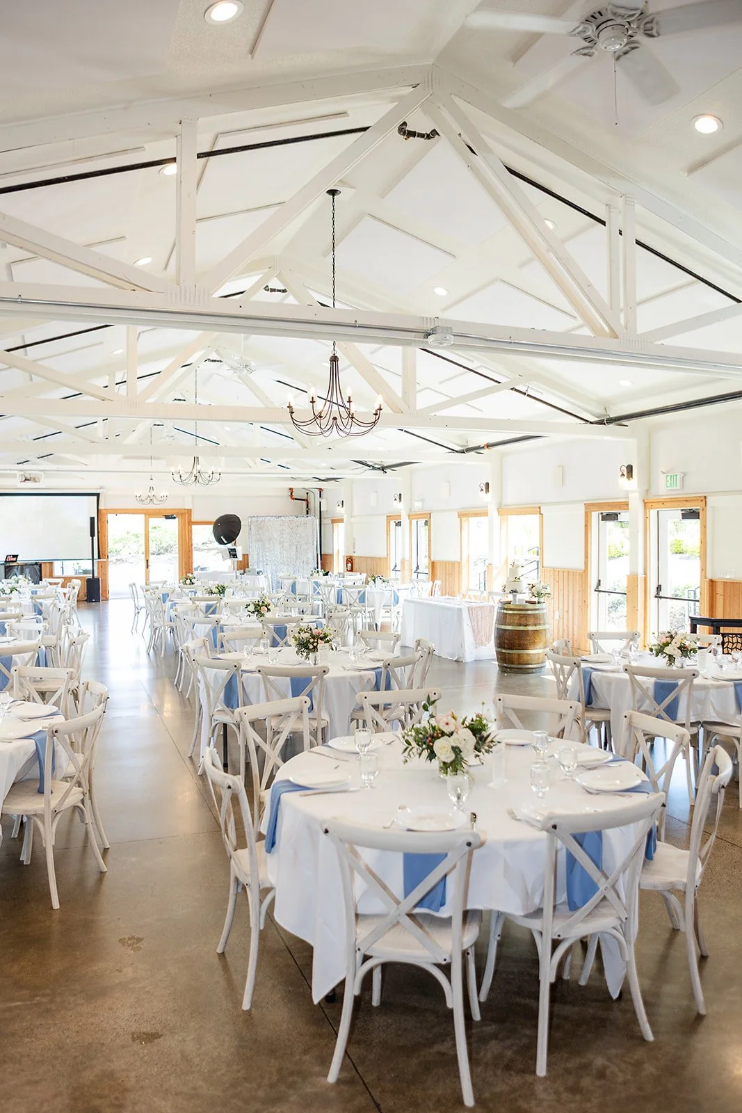 Event hall decorated for a celebration with round tables covered in white tablecloths, each adorned with floral centerpieces, white chairs, and set with plates, glasses, and napkins. The hall has a high, white, exposed-beam ceiling, chandeliers, and 