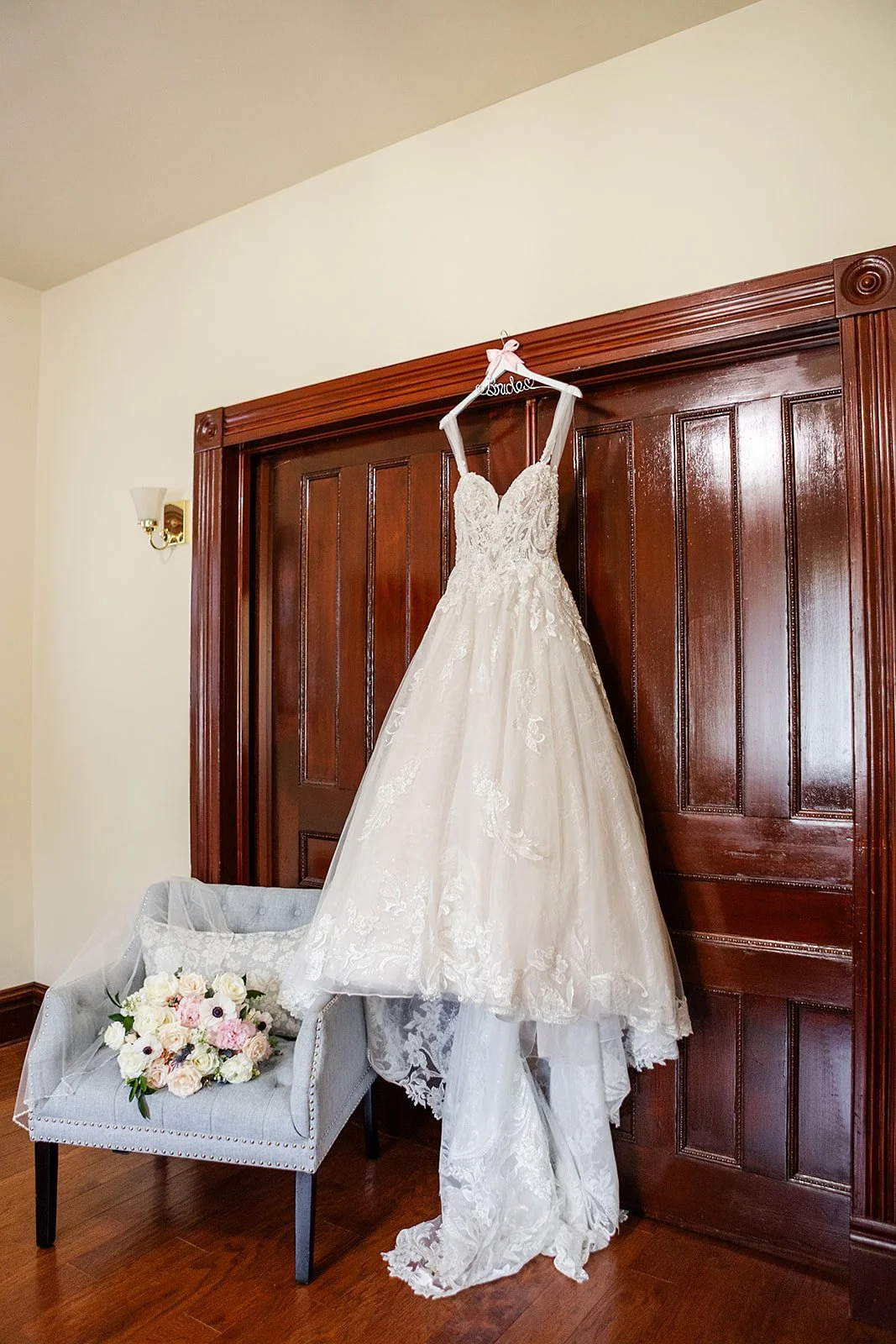 Bridal wedding dress hanging on curtain rod with a bouquet of flowers on a small armchair below.
